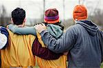 View from behind of three friends with their arms around one another in sports kit and woolly hats.