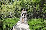 Active senior couple and dog crossing footbridge among lush green trees