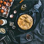 scrambled eggs or omelette in cast-iron frying pan and kitchen accessories top view. Eggs,shell,milk,parmesan cheese,scented rosemary salt,olive oil,pepper,apron,whisk,vintage knife on dark background