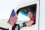 Caucasian man truck driver in the cab of his truck with an American Flag attached to the window.