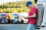 Caucasian man truck driver texting while standing next to the grill of his commercial truck in a struck stop.