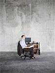 Business man sitting at a small desk in a large empty raw office space.