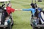 A senior couple doing a fist bump between golf carts on the fairway of a golf course.