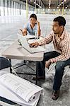 Asian woman and black man sitting at a desk in front of loading dock doors working on plans for new warehouse interior.