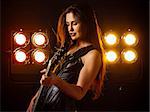 Photo of a beautiful young woman playing an electric guitar in front of stage lights.