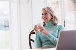 Smiling mature female freelancer drinking tea and working at laptop at home