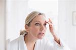 Mature woman touching eyebrow at bathroom mirror