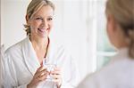 Smiling, confident mature woman in bathrobe drinking water at bathroom mirror