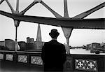 Rear view of man wearing black coat and Bowler hat standing on Tower Bridge over the River Thames in London, England, City Hall and the Shard skyscraper in the distance.