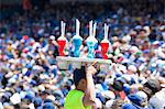 Side view of man carrying tray with soft drink bottles on his head, crowd of spectators in the background.