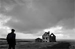 Rear view of man standing in landscape with ruin of stone building under a cloudy sky, ocean in the distance.