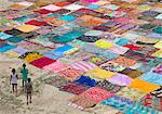 High angle view of three children standing on a field with a large number of colourful cotton fabrics spread out for drying.