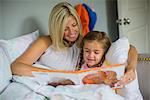 Mid adult woman in bed with daughter reading book