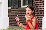 Young girl outdoors, holding small American flag, eating slice of watermelon