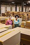 Team portrait of two African American female warehouse workers surrounded by products stored in cardboard boxes in front of loading dock doors in a large distribution warehouse.