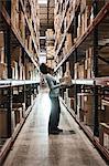 African American female warehouse worker checking inventory and carrying a box in an aisle of products stored in cardboard boxes, on large racks in a large warehouse distribution centre.