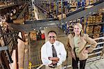 Overhead view portrait of a male Hispanic American executive in a shirt and tie and a Caucasian female executive in a jacket and slacks surrounded by large racks, forklifts and products stored in cardboard boxes  in a large distribution warehouse.