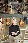 Overhead view portrait of a female Caucasian executive in a sweater and slacks surrounded by large racks, conveyor belts, forklifts and products stored in cardboard boxes  in a large distribution warehouse.