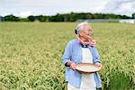Smiling elderly woman with grey hair standing in a rice field, holding bowl with freshly harvested rice grains.