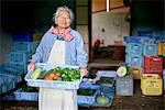 Elderly woman with grey hair standing in front of barn, holding blue plastic crate with fresh vegetables, smiling at camera.