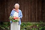Elderly woman with grey hair standing in a garden, holding a basket with fresh vegetables, smiling at camera.