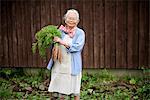 Elderly woman with grey hair standing in a garden, holding bunch of fresh carrots, smiling at camera.