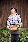 Woman with black hair wearing checkered shirt standing in a garden, holding basket with fresh vegetables, smiling at camera.