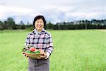 Woman with black hair wearing checkered shirt standing in a field, holding basket with fresh vegetables, smiling at camera.
