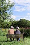 Husband and wife, rear view of elderly man wearing hat and woman sitting side by side on a bench in a garden.