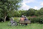 Husband and wife, rear view of elderly man wearing hat and woman sitting side by side on a bench in a garden with wheelchair parked alongside.