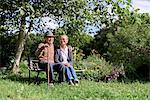 Husband and wife, elderly man wearing hat and woman sitting side by side on a bench in a garden.
