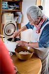 Elderly woman standing at a table in a kitchen, spooning sushi rice from rice cooker into bowl.