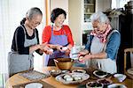 Three older women standing round a table in a kitchen, making sushi.