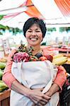 Smiling woman standing in a food and vegetable market, holding shopping bag with fresh produce including bananas, tomatoes and cabbage.