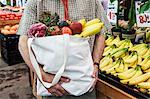 Close up of person at a food and vegetable market, holding shopping bag with fresh produce including bananas, tomatoes and cabbage.