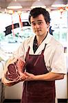 Man with brown curly hair wearing apron standing in butcher shop, holding beef forerib, smiling at camera.