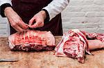 Close up of butcher wearing apron standing at a wooden butcher's block, preparing a rolled pork belly using string.
