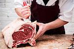 Close up of butcher wearing apron standing at a wooden butcher's block, butchering beef forerib.