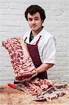 Man with brown curly hair wearing apron standing at a wooden butcher's block, holding beef forerib, looking at camera.