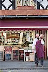 Man wearing apron standing on pavement outside butcher shop, carrying large piece of beef on his shoulder.