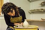 Woman with brown curly hair wearing apron shaping clay vase on pottery wheel.