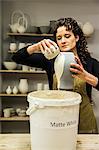 Woman with curly brown hair wearing apron standing in pottery workshop, pouring white glaze over unfired vase.