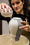 Woman with curly brown hair wearing apron standing in pottery workshop, pouring white glaze over unfired vase.