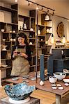 Woman with curly brown hair wearing apron standing in her pottery shop, looking at mobile phone.