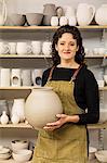 Woman with curly brown hair wearing apron holding unfired spherical clay vase.