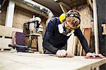 Man wearing ear protectors, protective goggles and dust mask standing in a warehouse, working on a piece of wood.
