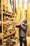 Man wearing ear protectors and dust mask standing in a warehouse, carrying long planks of wood on his shoulder.