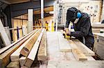 Man wearing ear protectors, protective goggles and dust mask standing in a warehouse, sanding planks of recycled wood.