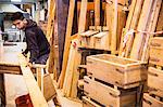 Young man wearing work gloves standing in warehouse, balancing plank of wooden on trestle, stack of recycled wooden crates.