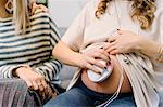 Pregnant woman and friend on sofa using prenatal listening device, cropped